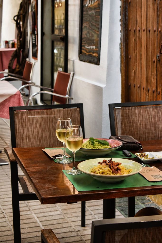 Mesa en la terraza de La Trattoria de Tarifa con dos platos de pasta y copas de vino blanco